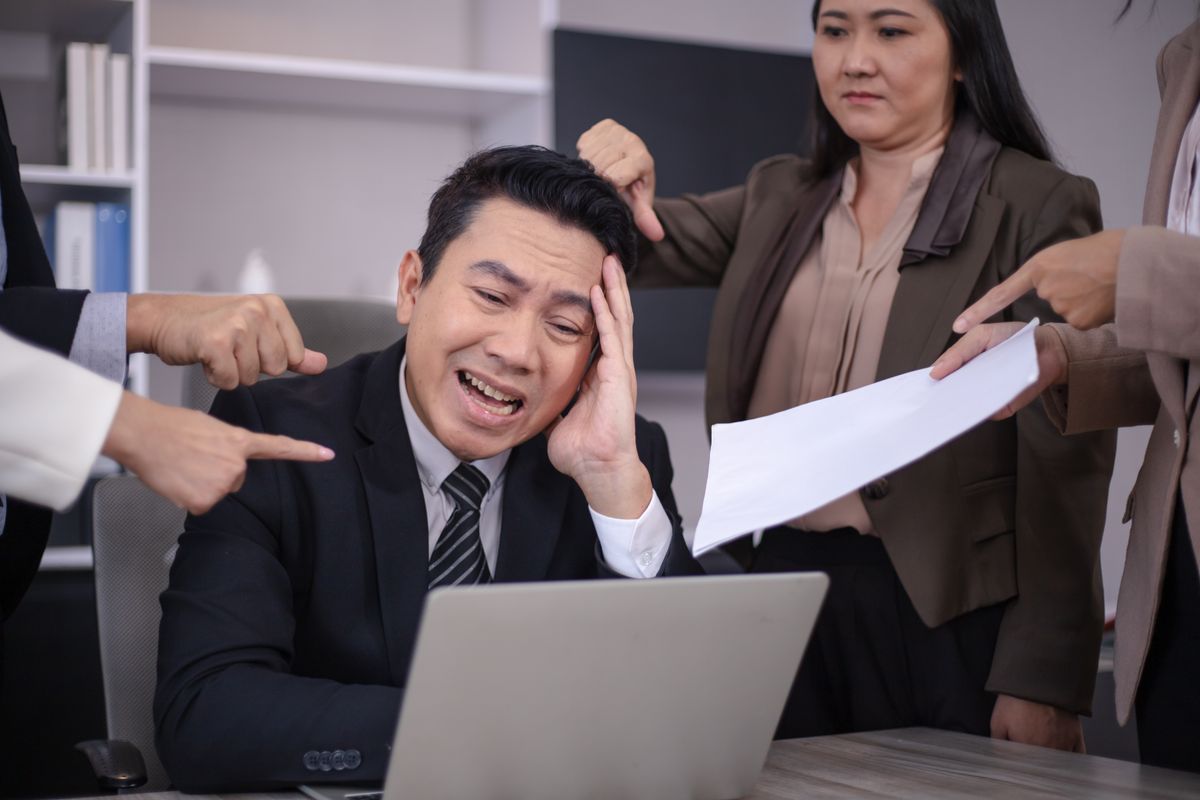 A stressed businessman at a laptop surrounded by colleagues pointing fingers