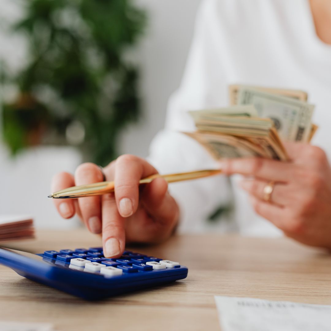 Person using a blue calculator and holding cash while planning a household budget at a desk