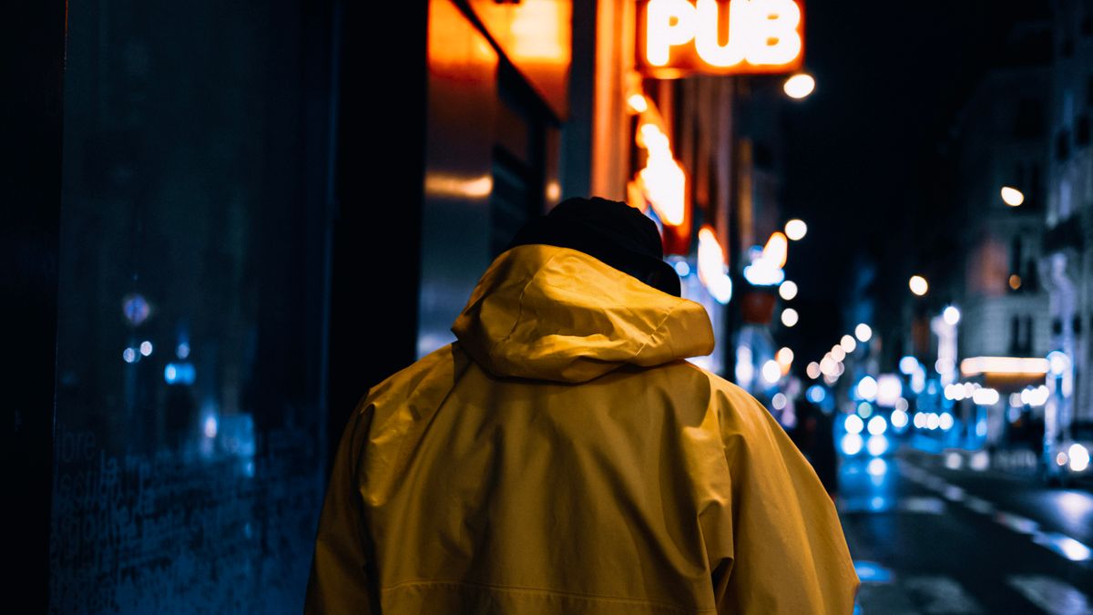A person in a yellow raincoat walking down a neon-lit city street at night