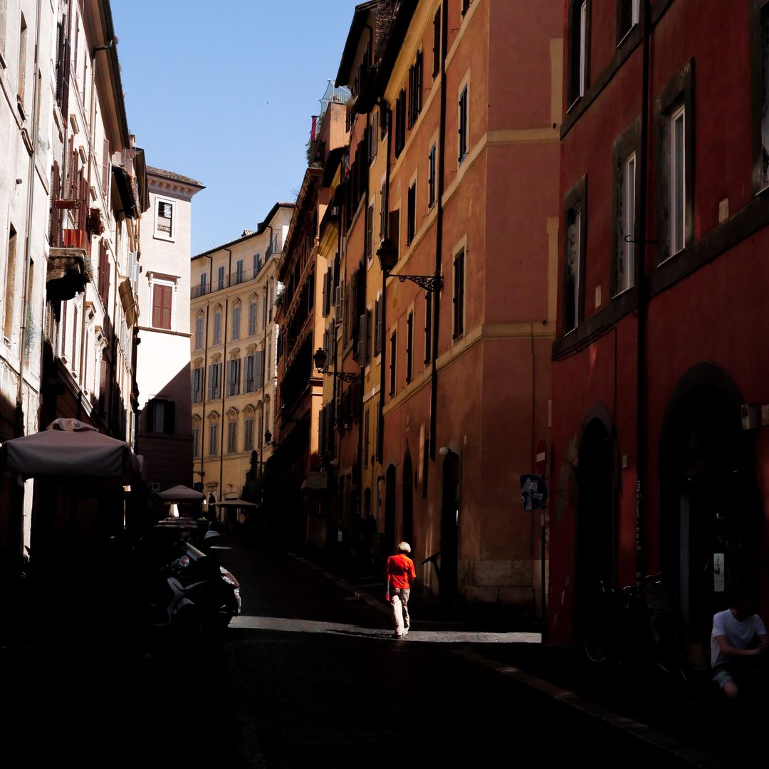 A person in a red jacket walking down a narrow sunlit old European street